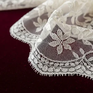 Detalle de blonda de seda Chantilly color crema, con patrón floral y brillo lustroso, sobre terciopelo. Ideal para vestidos de novia y alta costura.