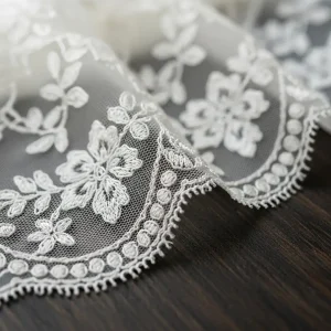 Primer plano de blonda Chantilly marfil, mostrando su delicado patrón floral y textura etérea sobre madera oscura. Ideal para vestidos de novia y lencería.