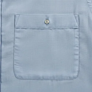Bolsillo de parche cuadrado de lino azul claro cosido en una camisa, mostrando la textura de la tela y costuras precisas. Detalle de confección.