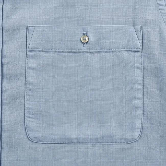 Bolsillo de parche cuadrado de lino azul claro cosido en una camisa, mostrando la textura de la tela y costuras precisas. Detalle de confección.