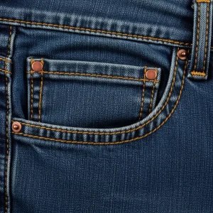 Primer plano del bolsillo delantero de un jean azul, mostrando el bolsillo francés y el bolsillo cerillero con costuras ocre y remache de cobre. Detalle de confección.