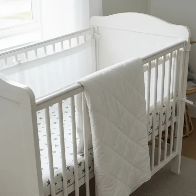 Cuna de bebé blanca vestida con ropa de cuna de algodón, incluyendo una sábana estampada y un edredón acolchado. Entorno de descanso seguro y confortable.