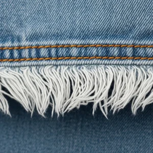 Borde deshilachado del bajo de un pantalón jean de denim, mostrando los hilos blancos sueltos. Acabado decorativo para un look casual.