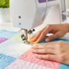 Manos guiando telas de patchwork bajo una máquina de coser, mostrando cómo acolchar una tela a máquina.